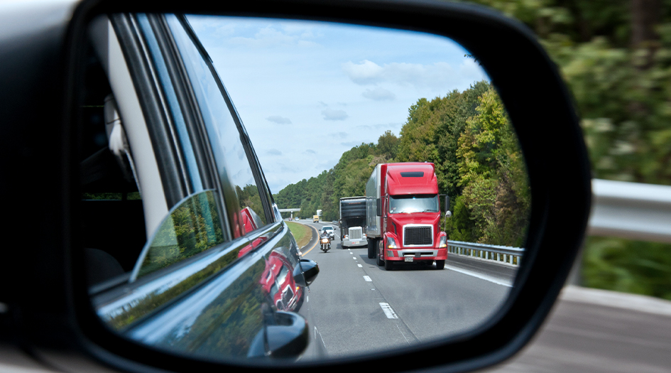 A close up of a side mirror shows a transport truck down the road, showing you how to safely share the road with transport trucks