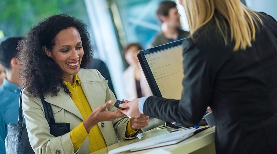 A woman accepts keys to her rental car from another woman behind a desk, after she learned how to save on rental vehicles