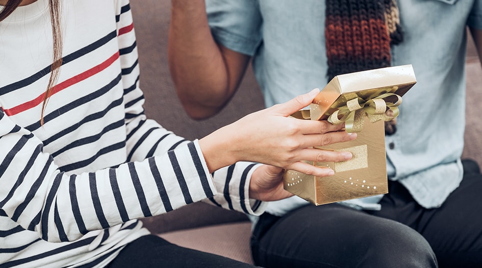 A woman hands an open gold present to a man, reminding you to insure your big-ticket Christmas gifts