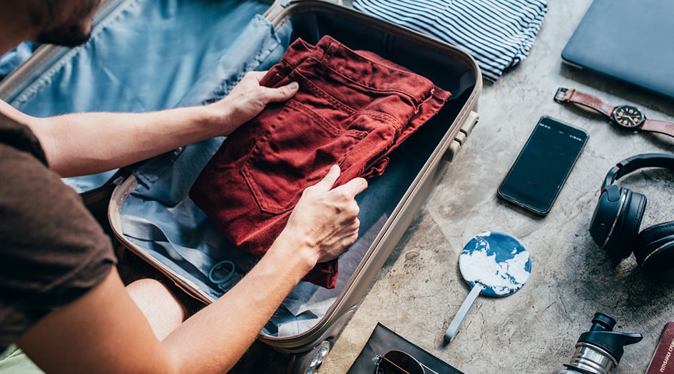 A man packs pants into a suitcase with various items nearby, as he follows a to-do list to prepare for a vacation