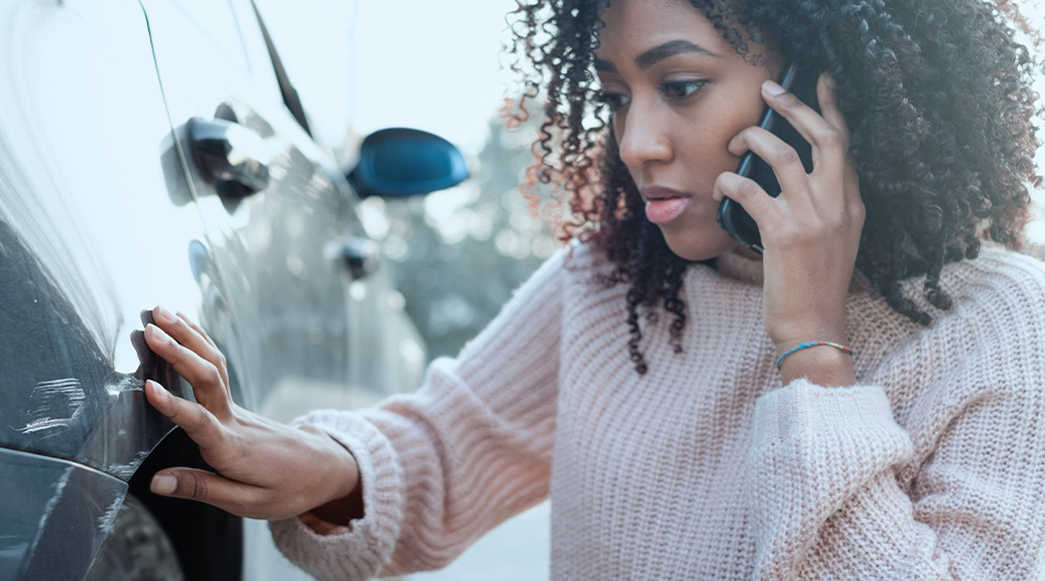 A woman is on the phone while she inspects damage to her car, showing one step to take immediately after a car accident.