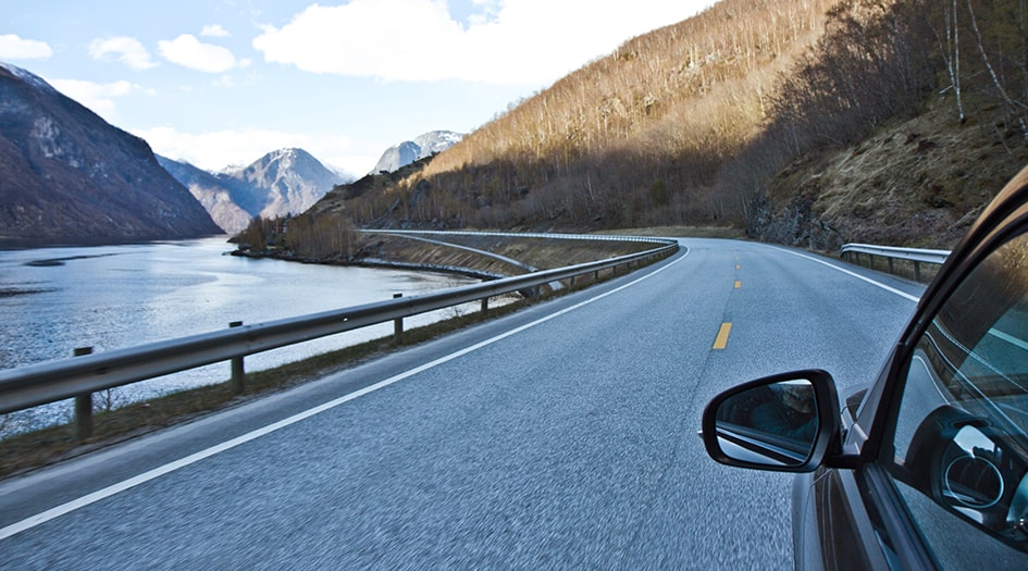 Consider whether to purchase public or private car insurance, as shown by the side of a car as it drives beside a lake and mountains