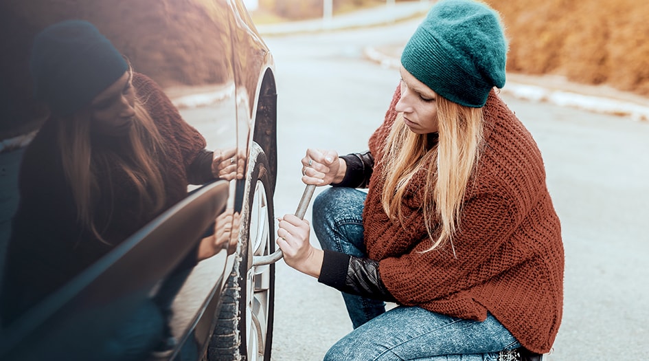 Une femme accroupie serre les boulons d’une roue de sa voiture, ce qui vous montre comment gérer à un éclatement de pneu.