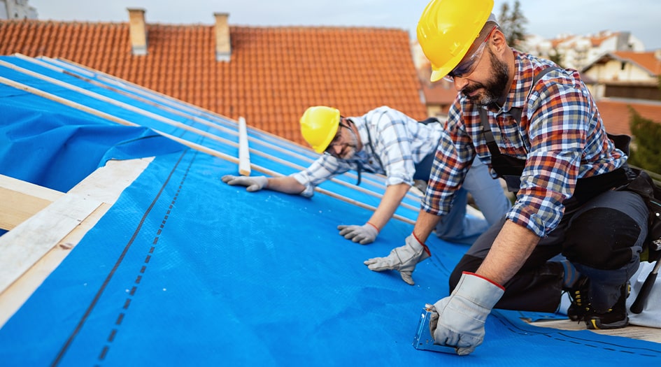 Two men staple blue material down before laying new roofing, showing one way to make your home more climate-resilient