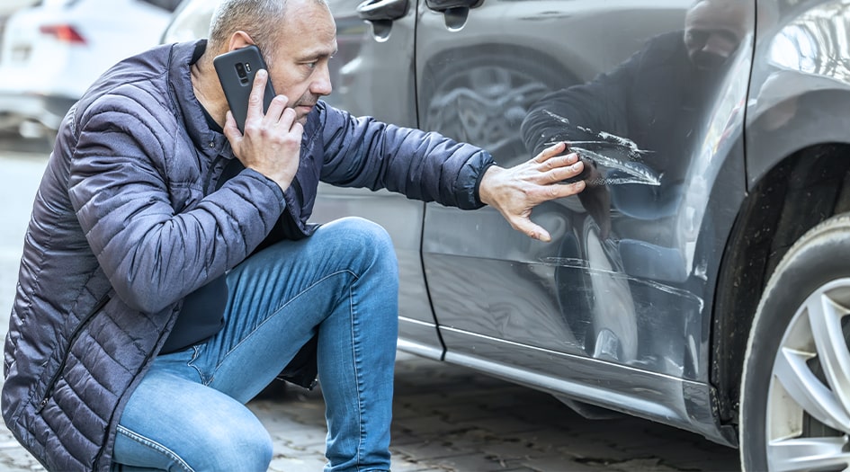 Un homme assis regarde une égratignure sur sa voiture et discute avec son assureur à propos du retrait d’une couverture.