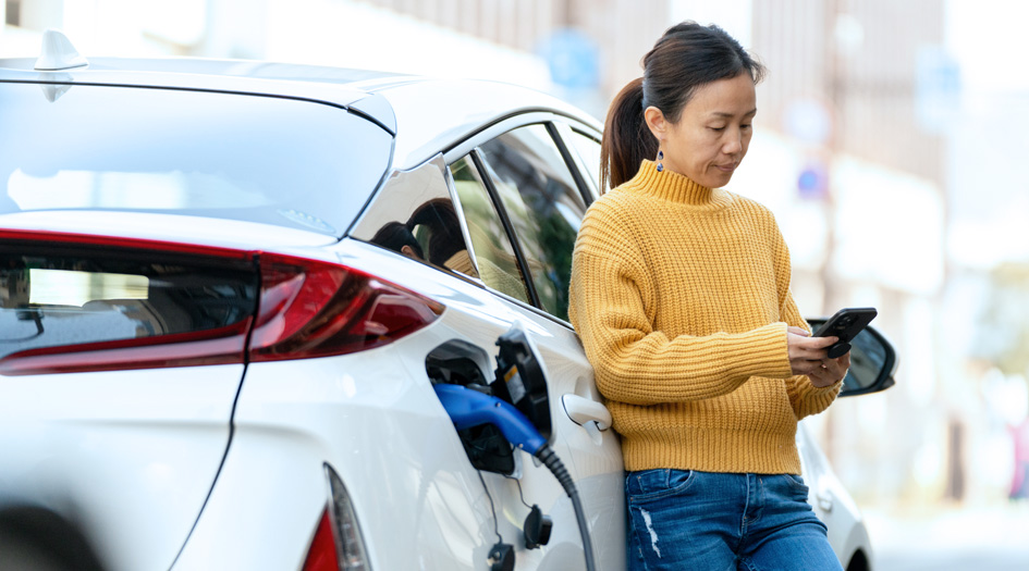 A woman reads about the pros and cons of electric and hybrid cars on her phone while her white car charges