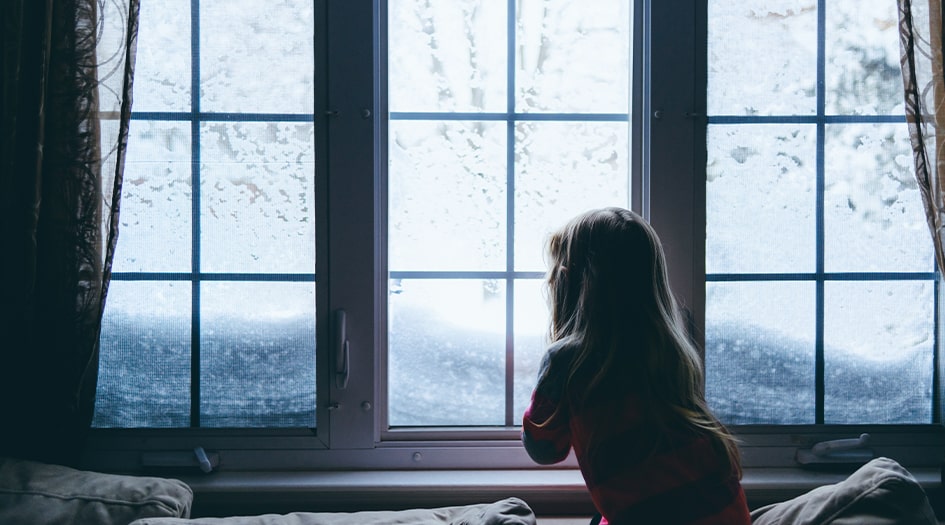 A child looks out a window as it snows, showing what to do before, during, and after a snowstorm