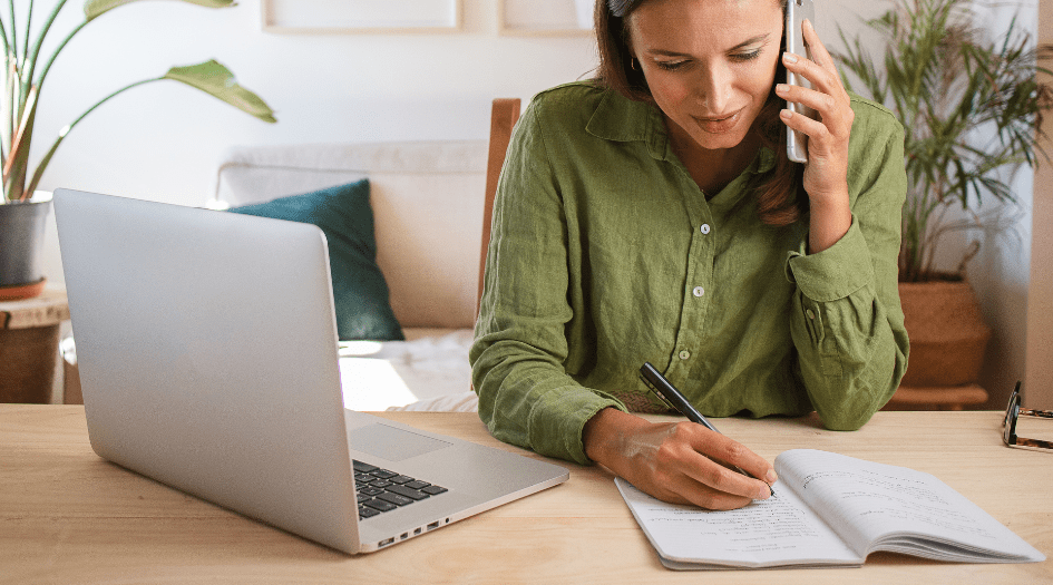 A woman is writing notes on a piece of paper as she shares information needed to file a home insurance claim over the phone.