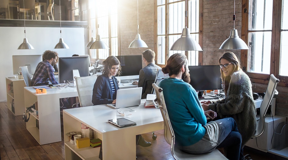 Six people work at a row of desks in an open-concept office, showing why it may be a good idea to buy group insurance over personal insurance