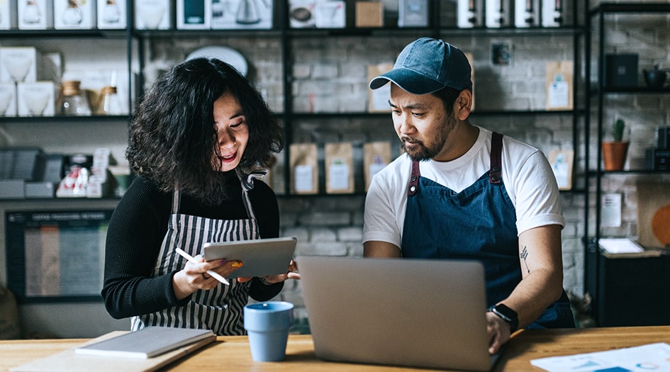 A man and woman sit in their small business and read why they need commercial liability insurance on a tablet and laptop