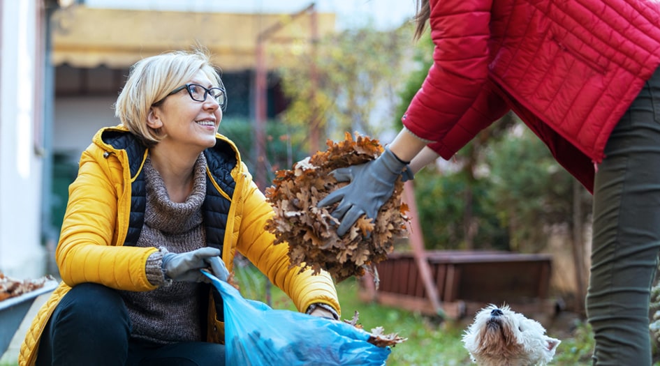 A woman holds a garbage bag open while another individual fills it with dead leaves, as part of the fall home maintenance checklist