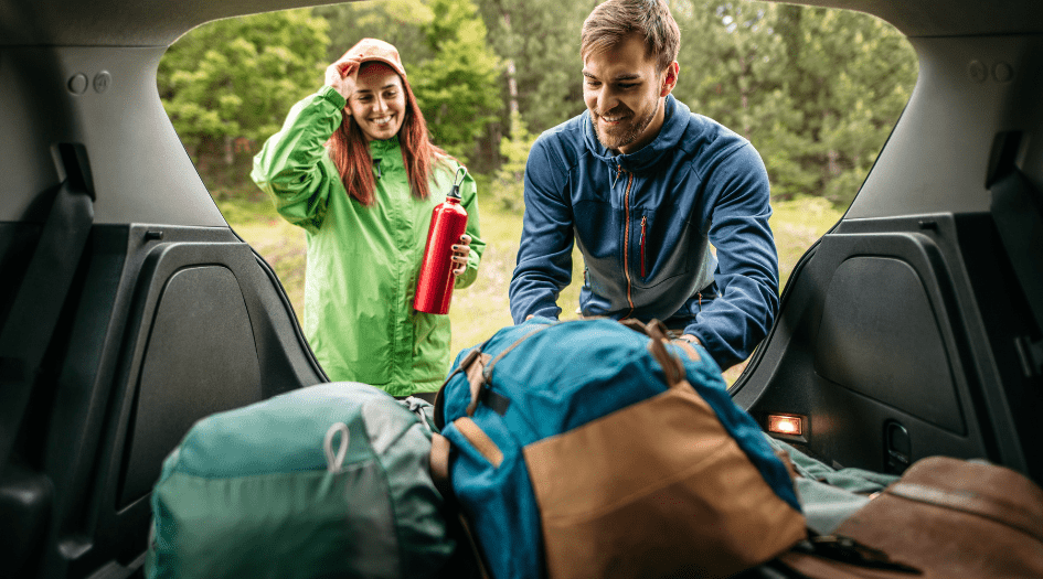 A man and woman pack the back of their car with multiple backpacks, showing how to pack for a safe fall camping trip.