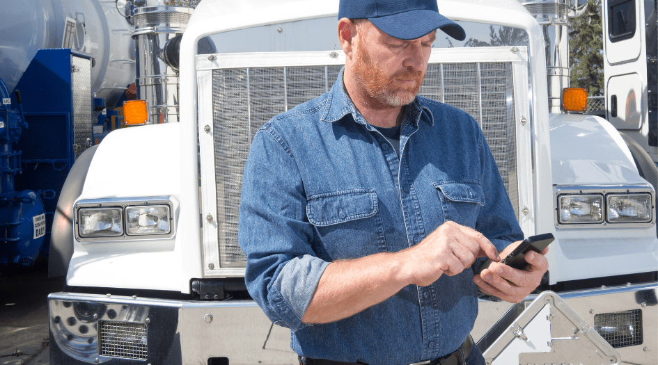 A man is in front of a transport truck typing on his phone, showing one step to filing a commercial car insurance claim.