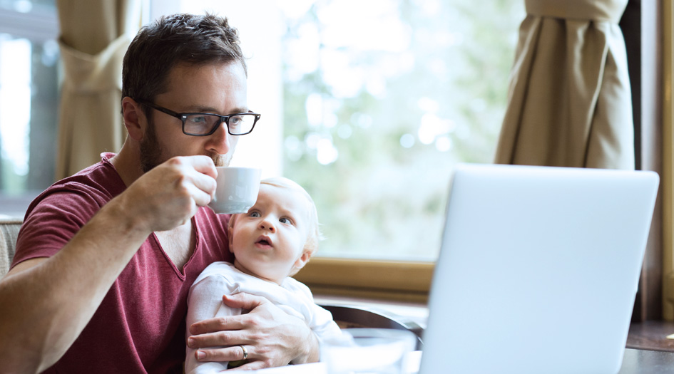 A man drinks coffee and reads about the common car insurance terms on his laptop while a baby sits on his lap.