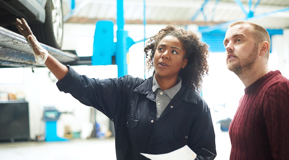 Une femme montre à un homme des dommages sur son véhicule dans un atelier de réparation, ce qui montre pourquoi vous devez comprendre les réclamations auto et les frais d’amélioration.